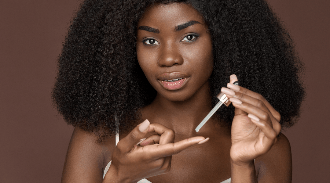 Best oils for low porosity hair growth: A Black woman with voluminous, healthy curls applying hair oil with a dropper. Using lightweight, nourishing oils is essential for moisturizing and strengthening low porosity hair.